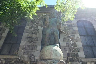 Statue eines kleinen Jungen mit einem Fisch in jedem Arm, stehend auf einem Sockel und umgeben von Bäumen.