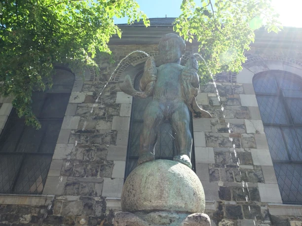 Fischpüddelchen Statue eines kleinen Jungen mit einem Fisch in jedem Arm, stehend auf einem Sockel und umgeben von Bäumen.