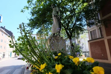 Wehrhafter Schmied Hinter einem Blumenkasten steht eine Bronze-Figur auf einem steinernen Sockel unter einem Baum.