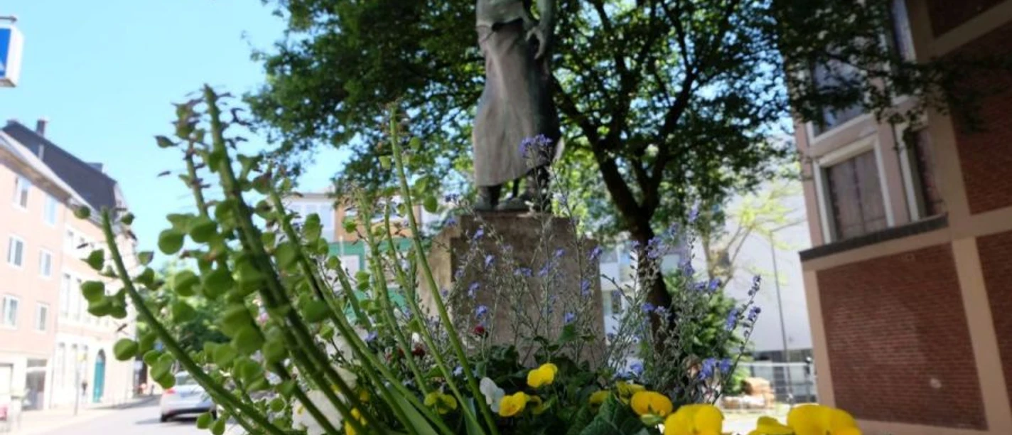 Wehrhafter Schmied Hinter einem Blumenkasten steht eine Bronze-Figur auf einem steinernen Sockel unter einem Baum.