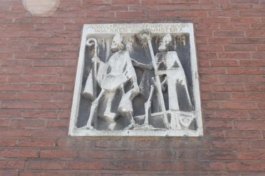 Mundolph und Gundolph Historisches Steinbild an einer roten Ziegelsteinwand mit zwei Figuren: Links ein Mann mit einem Hirtenstab und rechts eine skelettartige Gestalt, die einen Stab hält.