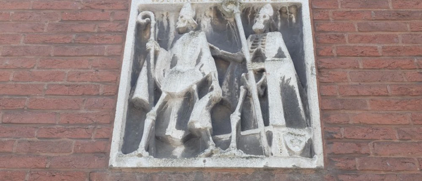 Mundolph und Gundolph Historisches Steinbild an einer roten Ziegelsteinwand mit zwei Figuren: Links ein Mann mit einem Hirtenstab und rechts eine skelettartige Gestalt, die einen Stab hält.