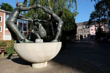 Eine Skulptur zeigt drei bronzene, lachende Figuren, die sich an den Händen halten und ein sitzendes Kind umgeben. Die Figuren stehen auf einem halbrunden Sockel vor einem Hintergrund aus Bäumen und Wohnhäusern.