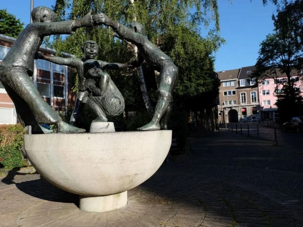 Eine Skulptur zeigt drei bronzene, lachende Figuren, die sich an den Händen halten und ein sitzendes Kind umgeben. Die Figuren stehen auf einem halbrunden Sockel vor einem Hintergrund aus Bäumen und Wohnhäusern.