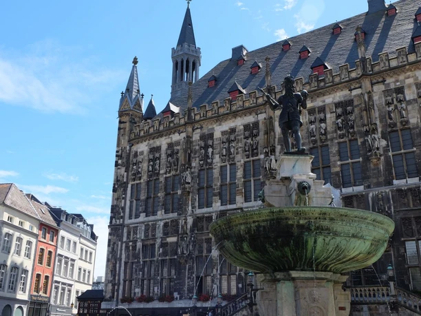 Karlsbrunnen Bronzestatue von Karl dem Großen auf einem Brunnen mit der historischen Fassade des Aachener Rathauses im Hintergrund.