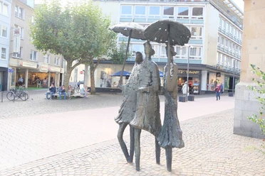 Bronzefiguren mit Regenschirmen an einer ruhigen Einkaufsstraße. Im Hintergrund verweilen mehrere Personen auf einer Bank unter einem Baum.