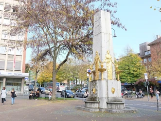 Hotmannspief Denkmal an einer befahrenen Straße mit zwei vergoldeten Statuen vor einem steinernen Obelisken, und Bäume mit gelben Herbstblättern. Menschen sind in der Nähe zu Fuß unterwegs.