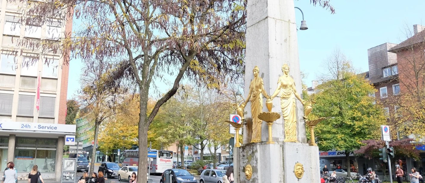 Hotmannspief Denkmal an einer befahrenen Straße mit zwei vergoldeten Statuen vor einem steinernen Obelisken, und Bäume mit gelben Herbstblättern. Menschen sind in der Nähe zu Fuß unterwegs.