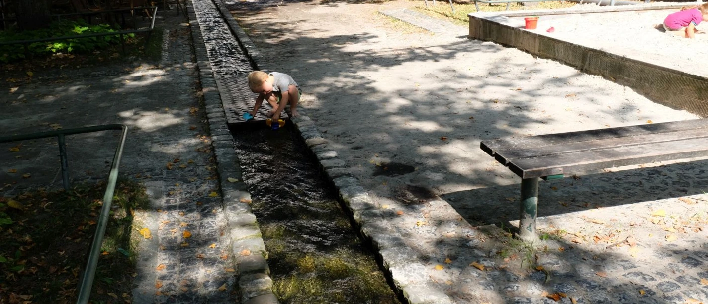 Ein kleines Kind spielt mit einem Förmchen an einem schmalen Bächlein im Schatten mehrerer Bäume. Rechts spielt ein kleines Mädchen in einem Sandkasten.