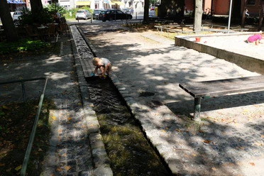 Johannisbach Ein kleines Kind spielt mit einem Förmchen an einem schmalen Bächlein im Schatten mehrerer Bäume. Rechts spielt ein kleines Mädchen in einem Sandkasten.