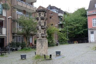 Kunstvoll bearbeitete Stele aus Stein steht auf gepflastertem Platz, umgeben von Bäumen und Wohngebäuden.