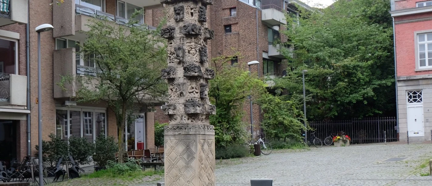 Stele am Augustinerplatz Kunstvoll bearbeitete Stele aus Stein steht auf gepflastertem Platz, umgeben von Bäumen und Wohngebäuden.