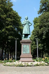Statue von einem Mann, der einen Arm in die Hüfte stützt, auf einem Sockel umgeben von Bäumen.