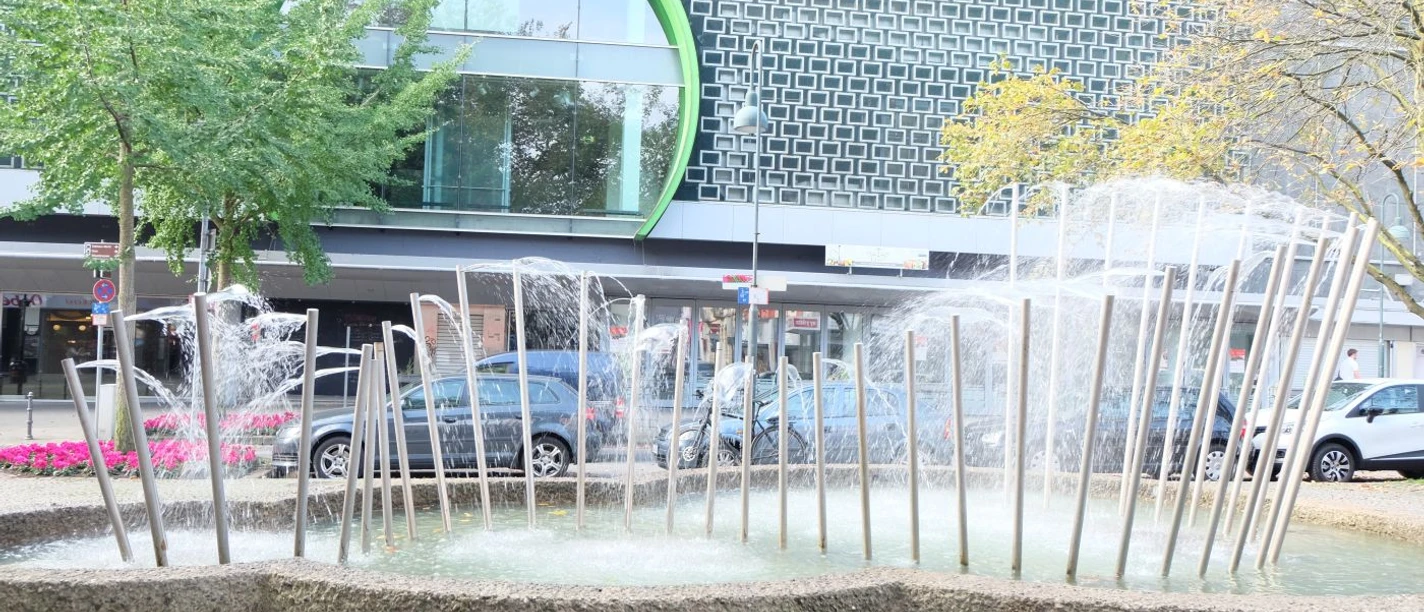 Brunnen mit einer Reihe Edelstahlröhren, aus denen Wasser spritzt, vor einem großen Gebäude.