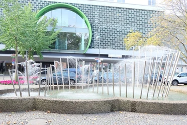 Röhrenbrunnen Brunnen mit einer Reihe Edelstahlröhren, aus denen Wasser spritzt, vor einem großen Gebäude.