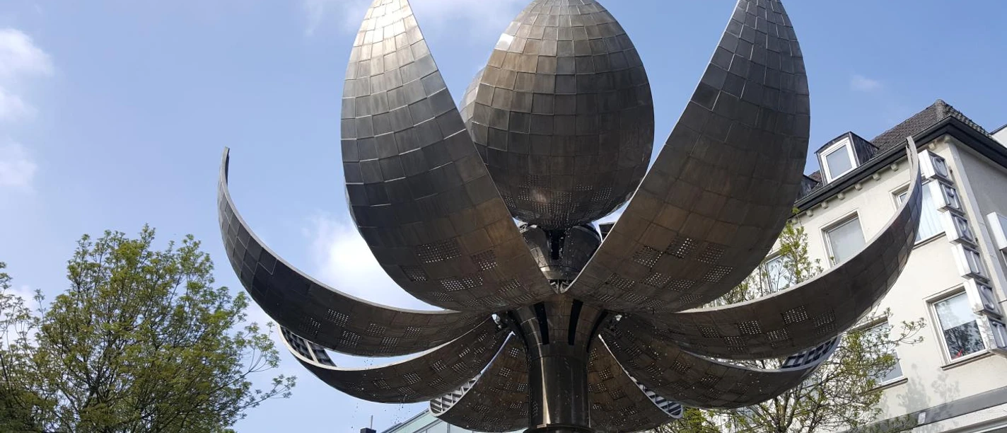 Metallskulptur in Form einer geöffneten Blüte, deren große Blütenblätter in den klaren Himmel ragen, umgeben von modernen Gebäudefassaden.