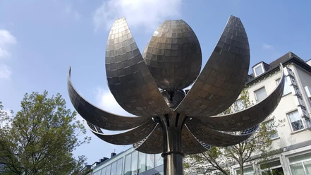 Kugelbrunnen Metallskulptur in Form einer geöffneten Blüte, deren große Blütenblätter in den klaren Himmel ragen, umgeben von modernen Gebäudefassaden.