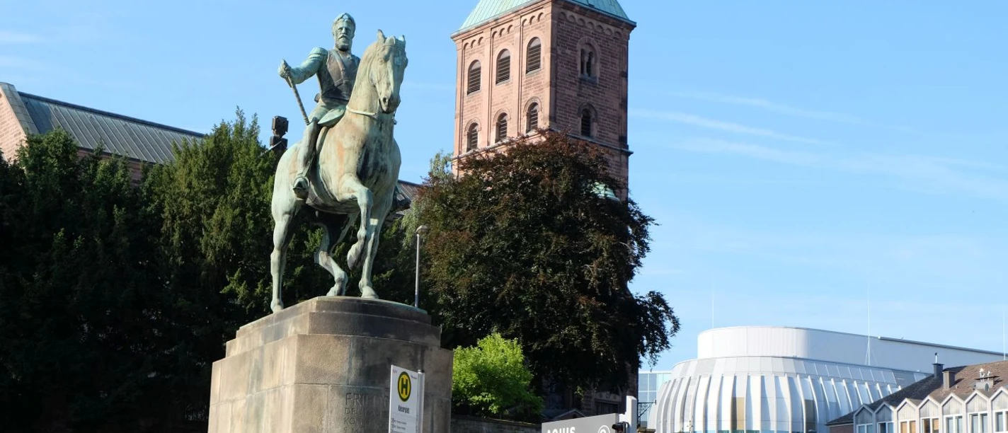 Reiterstandbild Kaiser Friedrichs III Auf einem steinernen Sockel steht das Denkmal von Kaiser Friedrich III auf einem Pferd. Im Hintergrund kann man das Dach des Einkaufszentrums Aquis Plaza erkennen.