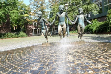 Drei fröhliche Kinder als Bronze-Skulpturen halten sich an den Händen und springen durch einen sprudelnden Brunnen.