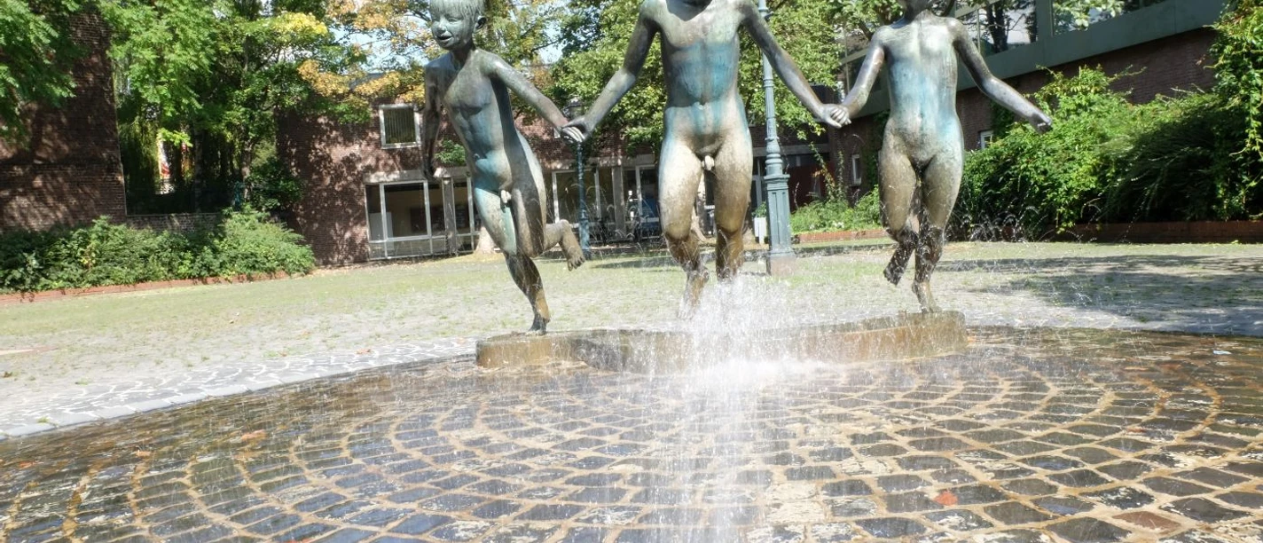 Laufende Kinder Drei fröhliche Kinder als Bronze-Skulpturen halten sich an den Händen und springen durch einen sprudelnden Brunnen.