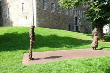 Zwei Figuren stehen einige Meter voneinander entfernt vor einem Wiesenstück und einem Hügel mit einem historischen Backstein-Gebäude dahinter.