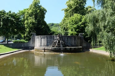 Teich mit halbkreisförmigem Brunnenbecken an seinem Ende, auf dem eine Skulptur aus Stein sitzt. Ringsherum stehen grüne Bäume.