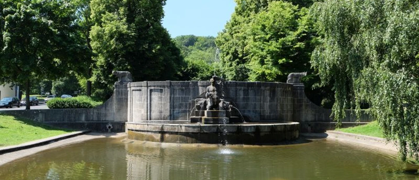 Teich mit halbkreisförmigem Brunnenbecken an seinem Ende, auf dem eine Skulptur aus Stein sitzt. Ringsherum stehen grüne Bäume.