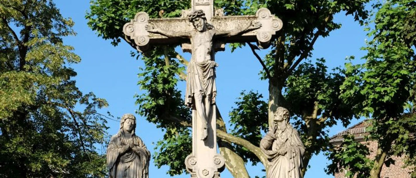 Steinernes Denkmal auf einem Sockel mit Christus am Kreuz zwischen zwei weiteren Steinfiguren, vor grünen Bäumen und strahlend blauem Himmel.