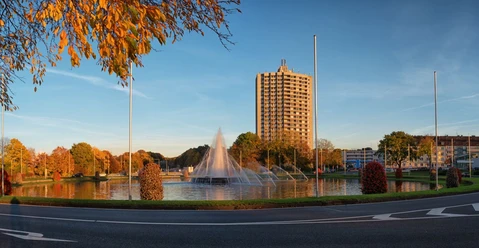 Großer, runder Brunnen mit Fontäne in der Mitte und einer Straße sowie Bebauung außen herum.