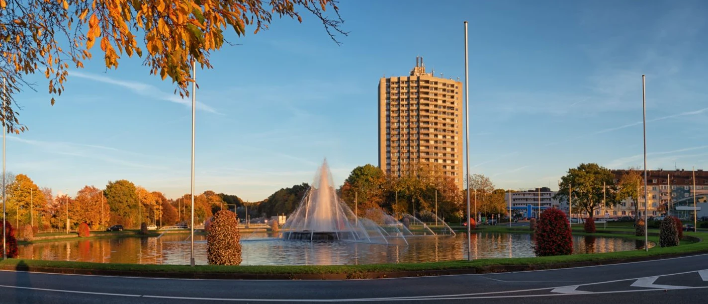 Europaplatz Großer, runder Brunnen mit Fontäne in der Mitte und einer Straße sowie Bebauung außen herum.