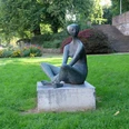 Eine Figur sitzt auf einem Sockel im Schneidersitz innerhalb einer Parkanlage.