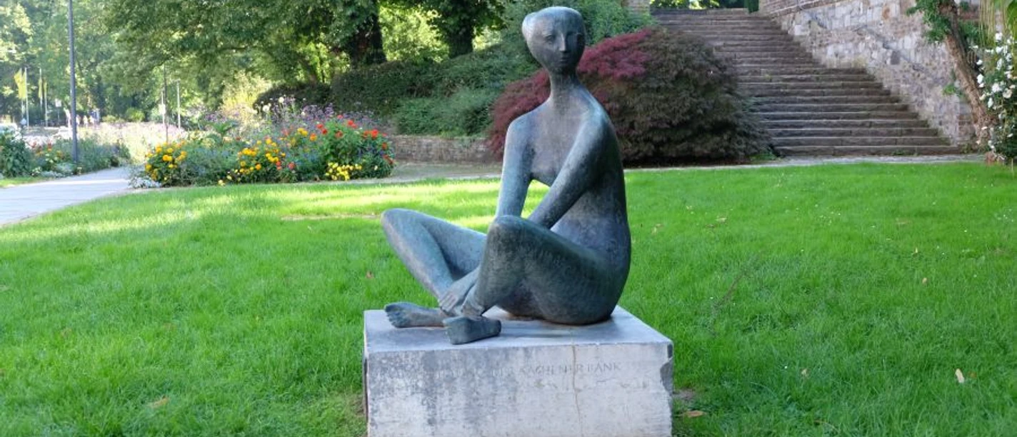 Eine Figur sitzt auf einem Sockel im Schneidersitz innerhalb einer Parkanlage.