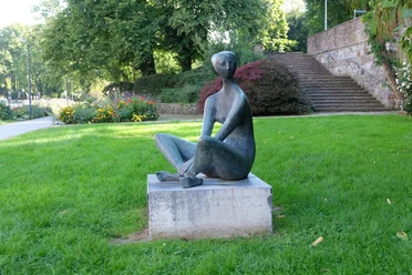 Die Sitzende Eine Figur sitzt auf einem Sockel im Schneidersitz innerhalb einer Parkanlage.
