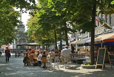 Belebter Marktplatz an einem sonnigen Tag. Menschen sitzen im Freien an Café- und Restauranttischen, während Bäume ihnen Schatten spenden.