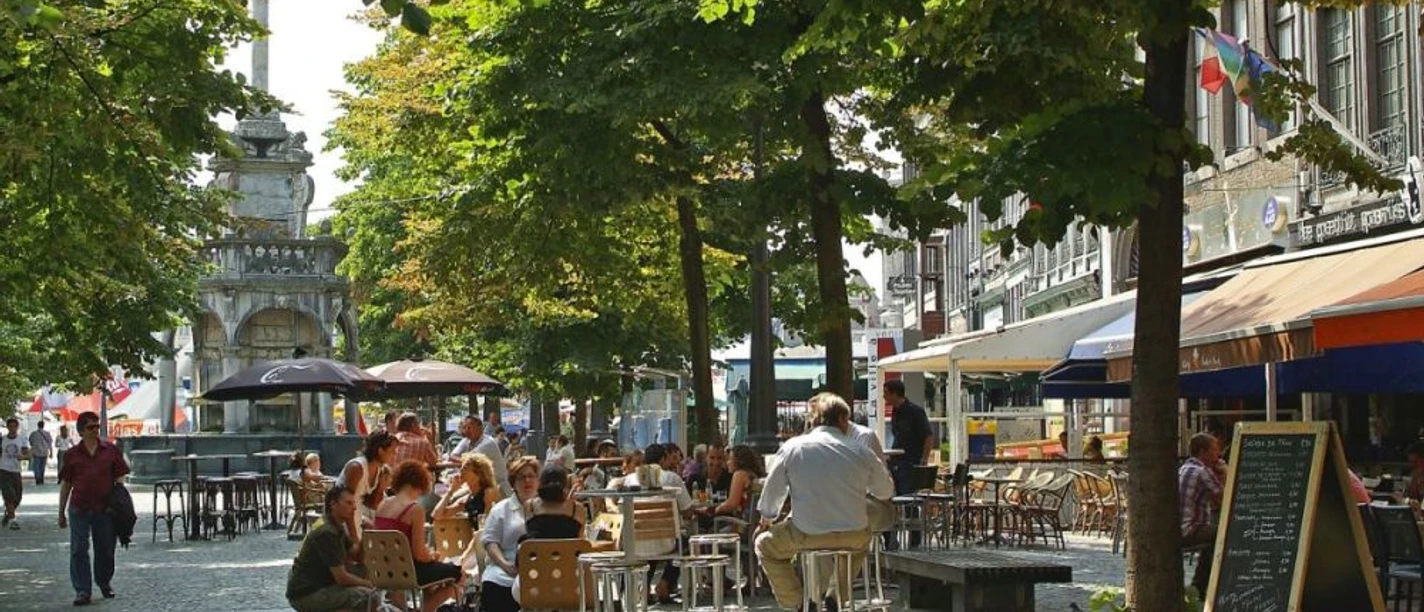 Lütticher Marktplatz Belebter Marktplatz an einem sonnigen Tag. Menschen sitzen im Freien an Café- und Restauranttischen, während Bäume ihnen Schatten spenden.