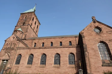 Kirche aus rotem Sandsteinbau, gotischen Fenstern und einem Turm unter klarem, blauem Himmel.