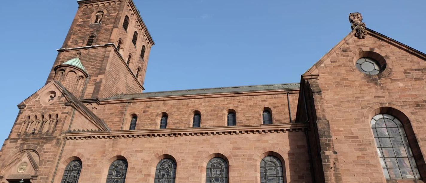 Kirche aus rotem Sandsteinbau, gotischen Fenstern und einem Turm unter klarem, blauem Himmel.