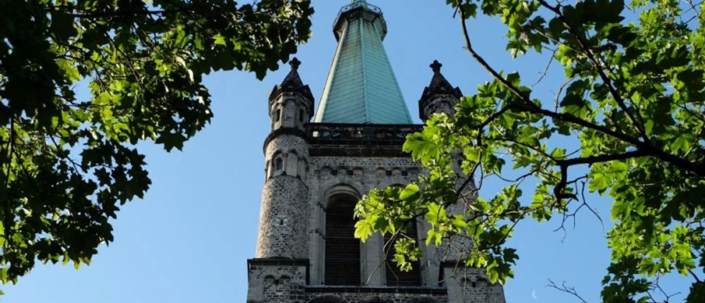 Hinter zwei Bäumen ragt ein Kirchturm mit Kirchturmspitze in den blauem Himmel.