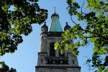 St. Jakob Hinter zwei Bäumen ragt ein Kirchturm mit Kirchturmspitze in den blauem Himmel.