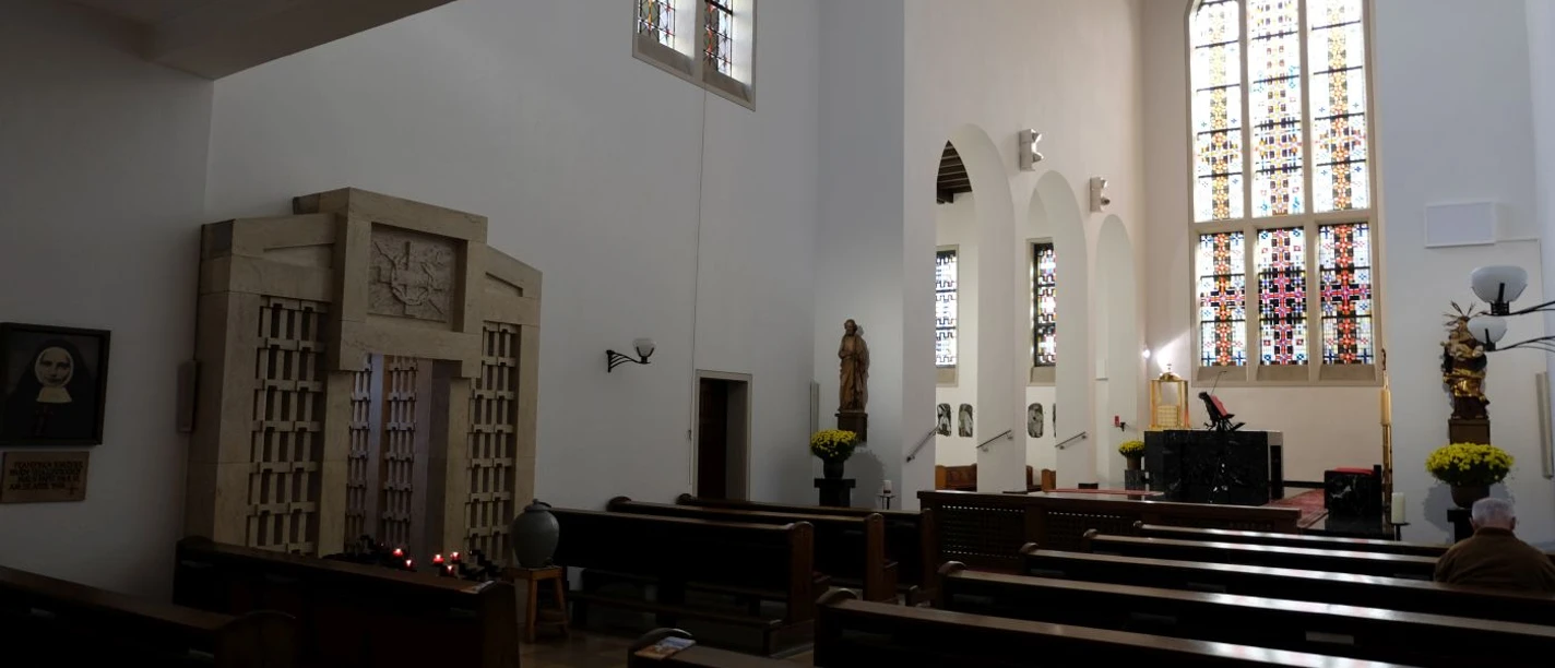 Innenansicht der Klosterkirche mit schlichter Einrichtung, Kirchenbänken und bunten Glasfenstern.