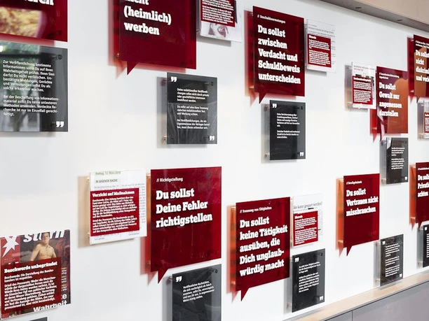 Museumswand mit mehreren Informationstafeln in einem klaren, roten und weißen Design. Die Tafeln zeigen verschiedene historische Zeitungsexzerpte mit markanten Zitaten und Bildern. Die Anordnung der Tafeln ist asymmetrisch.