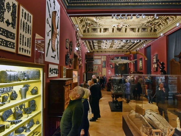 Besuchende in einem dunkelrot gestrichenen Museumsraum bewundern verschiedene Exponate, wie Krüge und Waffen, und Deckenverzierungen.