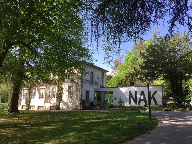 Ein Gebäude mit großen Fenstern zwischen dichtem, grünem Laub im Sonnenschein. Die weiße Mauer links trägt die großen Buchstaben "NAK".
