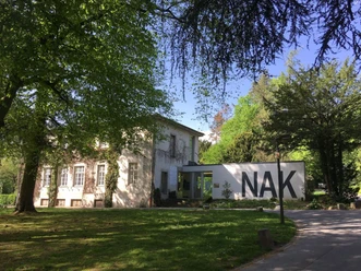Neuer Aachener Kunstverein Ein Gebäude mit großen Fenstern zwischen dichtem, grünem Laub im Sonnenschein. Die weiße Mauer links trägt die großen Buchstaben "NAK".
