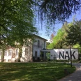 Neuer Aachener Kunstverein Ein Gebäude mit großen Fenstern zwischen dichtem, grünem Laub im Sonnenschein. Die weiße Mauer links trägt die großen Buchstaben "NAK".