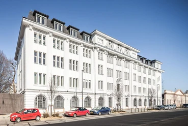 Stadtarchiv Aachen Helles, langes, historisches Gebäude mit vier Stockwerken und hohen Fenstern mit Sprossen. Vor dem Gebäude stehen einige parkende Autos.