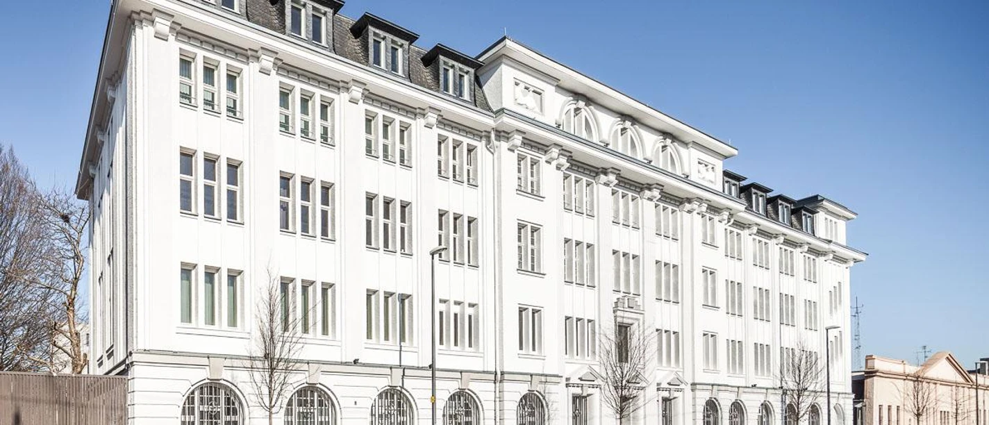 Stadtarchiv Aachen Helles, langes, historisches Gebäude mit vier Stockwerken und hohen Fenstern mit Sprossen. Vor dem Gebäude stehen einige parkende Autos.