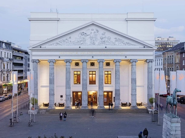 Weiße Fassade des Theater Aachen mit klassizistischen Säulen, Dachverzierungen und beleuchtetem Eingangsbereich in der Abenddämmerung.