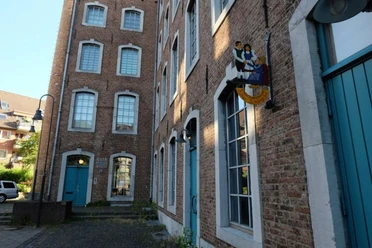 Backsteingebäude mit großen Fenstern und hellblauen Türen. An einer Fassade hängt ein buntes, kunstvolles Schild.