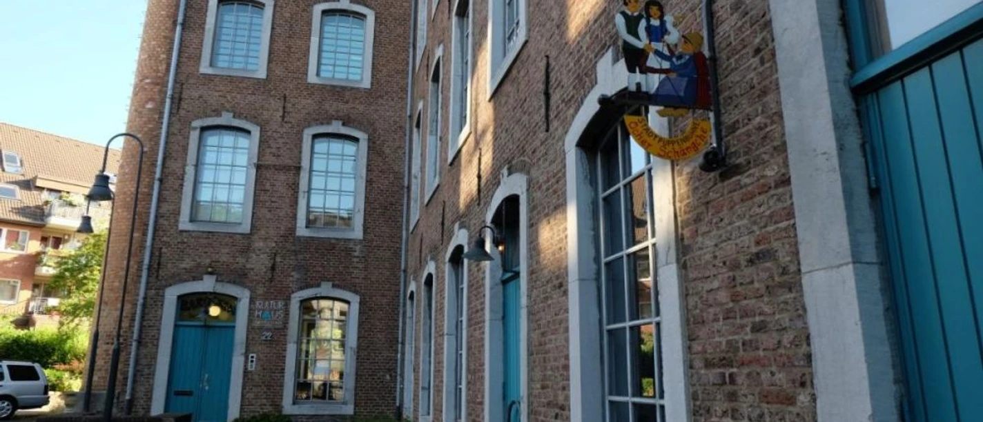 Backsteingebäude mit großen Fenstern und hellblauen Türen. An einer Fassade hängt ein buntes, kunstvolles Schild.